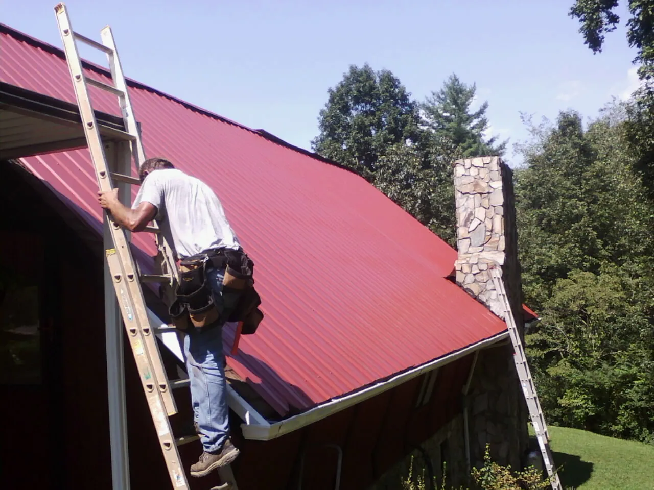 Roofer on a ladder beside a red metal roof and stone chimney — Soffit & Fascia Repair by Roofing Brothers in Days Creek
